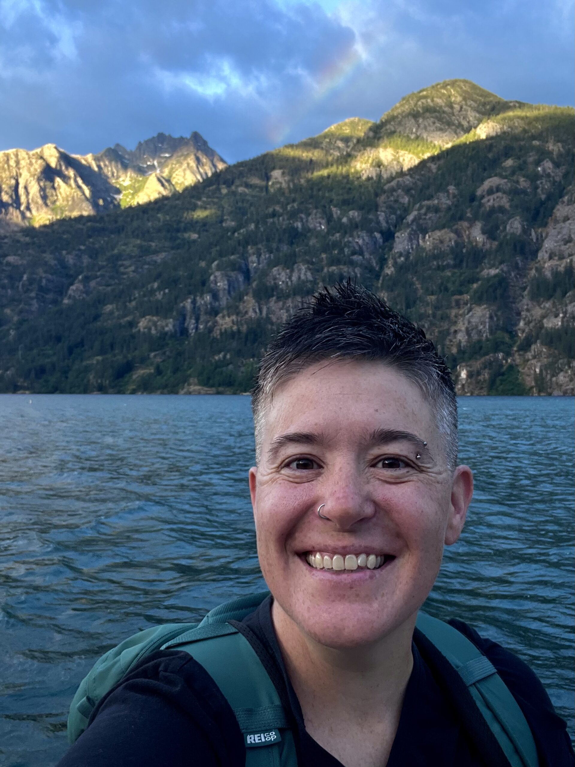 IMG_5390_Original Kris smiles at the camera while wearing a backpack. There are large mountains with a rainbow overhead in the background.